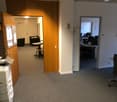 The image shows an office space with a carpeted floor, wooden doors, and various office equipment such as a printer and computer monitors. The room appears to have a waiting area with a chair and a small table.