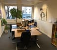 The image shows an office space with a large wooden desk, a computer, and various office supplies and equipment. There is a potted plant on the desk and artwork on the walls. The room has large windows overlooking the outdoors.
