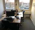 The image shows an office workspace with a desk, computer monitor, keyboard, phone, and other office equipment. The room has large windows overlooking a street outside with trees and buildings visible. The walls are light-colored and the floor appears to