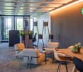 Modern and open office space with large windows, comfortable seating areas, potted plants, and a dining table. The space has a warm and inviting atmosphere with a mix of gray and orange tones.