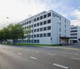 A multi-story office building with a modern, gray exterior and large windows. The building has multiple floors and appears to be located on a street with multiple lanes of traffic and pedestrian walkways.