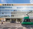A multi-story commercial building with a Credit Suisse bank branch and a Garage Keigel auto repair shop on the ground floor. The building has a glass facade with a grid-like pattern. There are people walking on the sidewalk in front of the building, and a