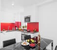 Modern white kitchen with red accent walls, built-in microwave, under-counter dishwasher, large dining table, high-gloss finish, stainless steel appliances, glass tabletop, wine bottle, glasses, fruit bowl
