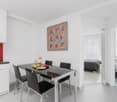 Modern dining area with glass table and black chairs, white kitchen cabinets, red kitchen wall, oven, microwave, dining table, wine bottle, glasses, and decorative items. There is a bedroom with a bed, pillows, and a painting in the distance.