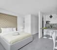 white interior, king size bed, 2 night tables, small kitchen, breakfast bar, 3 bar stools, round lamp, small plants, countertop vases