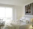 White walls, zebra painting, white bed with white bedding, bedside table with lamp, desk with stack of books, chair, large window with curtains, tiled floor