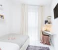 small room with single bed, desk, white shelves, picture frames, tv, large white curtains, pink chair cloth
