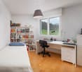 A small bedroom with a bed, a desk, a chair, a bookshelf, and a window with a view of a grassy field.