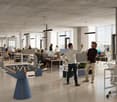 Large open-concept office space with concrete ceiling, modern lighting fixtures, and floor-to-ceiling windows providing a view. The space features a mix of workstations, lounge areas, and a foosball table. The office appears to have a casual, collaborativ