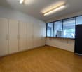This image shows a large, empty room with white walls and a wooden floor. The room has several built-in cabinets or storage units along one wall, and a large window that provides natural light. The room appears to be a multipurpose space, possibly used fo