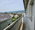 The image shows a balcony or terrace overlooking a residential neighborhood. The balcony has a railing and there are several houses and apartment buildings visible in the distance, with some trees and greenery in the foreground.