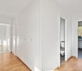 The image shows a hallway with white walls, wooden floors, and built-in closets along one side. There is a doorway leading to another room visible at the end of the hallway.