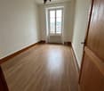 Spacious bedroom with hardwood flooring, white walls, and a large window providing a view of the surrounding area. The room features a chandelier lighting fixture.