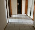 The image shows a hallway with white walls, a tiled floor, and wooden door frames. The hallway appears to be well-lit with a light fixture hanging from the ceiling.