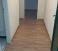 The image shows a hallway with white walls and a wooden floor. The hallway leads to a room with a blue wall visible through the open doorway.