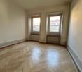 This image shows a room with a hardwood floor in a herringbone pattern. The room has two large windows that provide natural light. The walls are painted a neutral color. The room appears to be empty, with no furniture or decor visible.