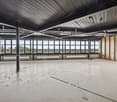 Large open space with high ceilings, concrete floor, and large windows offering a panoramic view of the surrounding area. The space appears to be a commercial or industrial building, possibly an office or warehouse.