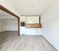 The image shows an open-plan living space with a kitchen area. The kitchen features white cabinets and drawers, a built-in oven, and a wooden backsplash. The flooring throughout the space is a light-colored wood-look laminate. There is a wooden beam visib