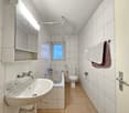Bathroom with white tiled walls and floor, bathtub, toilet, sink, mirror, towels on towel rack, power outlet, door.