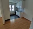 Small, empty kitchen with wooden floors, white kitchen units, dark countertops, white radiator, and window