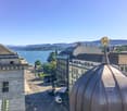 Exterior building view: Large modern buildings, golden dome on top, street with road and tram tracks, clear view of mountains and lake