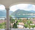Arched balcony with ornate wrought-iron railing, overlooking a scenic lake surrounded by mountains and lush greenery