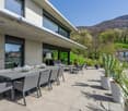 Modern concrete house with large windows, outdoor patio with dining table and lounge chairs, surrounded by lush greenery and mountains in the background