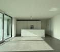 The image shows a spacious, open-concept living area with a modern kitchen. The room features large windows that provide ample natural light and offer a scenic view of the surrounding landscape. The kitchen is equipped with sleek, white cabinets and a cen