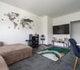 Modern living room with a large world map wall decor, a gray sectional sofa, a black entertainment unit with shelves and a TV, a green and white patterned rug, and a blue accent chair.