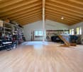 Large open space with wooden beamed ceiling, hardwood floors, shelving units with various items, musical instruments like a piano, and a fireplace visible in the corner.