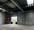 Empty warehouse with high ceiling, metal beams, large windows, storage shelves, ladder, and a door.