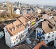 Aerial view of a row of terraced houses with brown roofs, white walls, and balconies. The houses are situated along a street with a river running through the town.