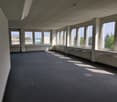 Large open office space with floor-to-ceiling windows, gray carpeted flooring, and white walls. The room appears to be empty and ready for use.