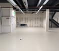 An empty room with white walls, white floor, exposed pipe and wires on ceiling, door, and stair