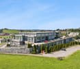 Multistory building, gray exterior, multiple windows, rooftop parking, stone wall enclosure, landscaped area, greenery, multiple cars parked