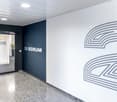 The image shows an office space with a dark blue wall featuring the text 'SCRUM 205' and a large graphic design on the opposite white wall. The room has a glass door entrance, a tiled floor, and some potted plants, indicating a professional and modern off