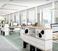 Spacious open-plan office with large windows offering a panoramic view of the city skyline. The office features modern workstations with computers, storage units, and ergonomic chairs. The overall design is minimalist and bright, creating a professional a