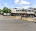 The image shows a busy street scene in an urban area. There are several buildings, including a cafe or shop, and people walking on the sidewalk. The street has yellow crosswalk markings and there are parked cars visible. The sky is partly cloudy.