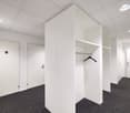 The image shows a modern office space with white walls, a carpeted floor, and recessed lighting. There are two doors visible, likely leading to other rooms or offices. The space features a built-in closet or storage unit with a clothes hanger visible insi