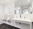 Large, modern bathroom with white tiled walls and floors, two white sinks with chrome faucets, and a dishwasher visible in the corner.