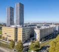 Two high-rise buildings with yellow and white exterior, large windows, and a modern architectural design. The buildings are surrounded by trees and a green lawn. In the distance, there are more buildings and a clear blue sky.