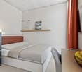 Bedroom with bed, pillows, blanket, a shelf with a picture frame and a grid, TV mounted on the wall, table with fruit bowl, and a vase with flowers