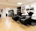 A hair salon interior with wooden floors, white walls, round lamps, black swivel chairs, shampoo trolleys, shelves with products, and framed pictures on the wall.