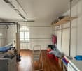 Large open space with wooden floors, white walls, a white washing machine, shelves, a ladder, red baskets and a blue hospital bed.