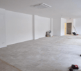 An empty room with white walls, grey tiled floor, air conditioner mounted on the ceiling, small items on the floor, and an electrical outlet.