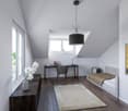 A room with a desk, chair, bed, and a painting on the wall. The room has a wooden floor, white walls, and a ceiling lamp. There is a vase with white flowers on a wooden table, and a rug on the floor. The desk has a laptop, books, and a lamp. The bed has a