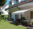 white house with brown roof, awning, terrace with outdoor dining set, garden with flowers and shrubs