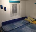 Blue double bed, yellow decorative pillows, white sheer curtains, wall mounted frames, wooden shelf with items
