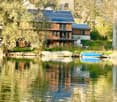 two-story wooden house, solar panels, trees, lake with boats