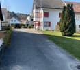 Driveway leading to a house with red shutters, brown roof, white walls, and a small garden in front.